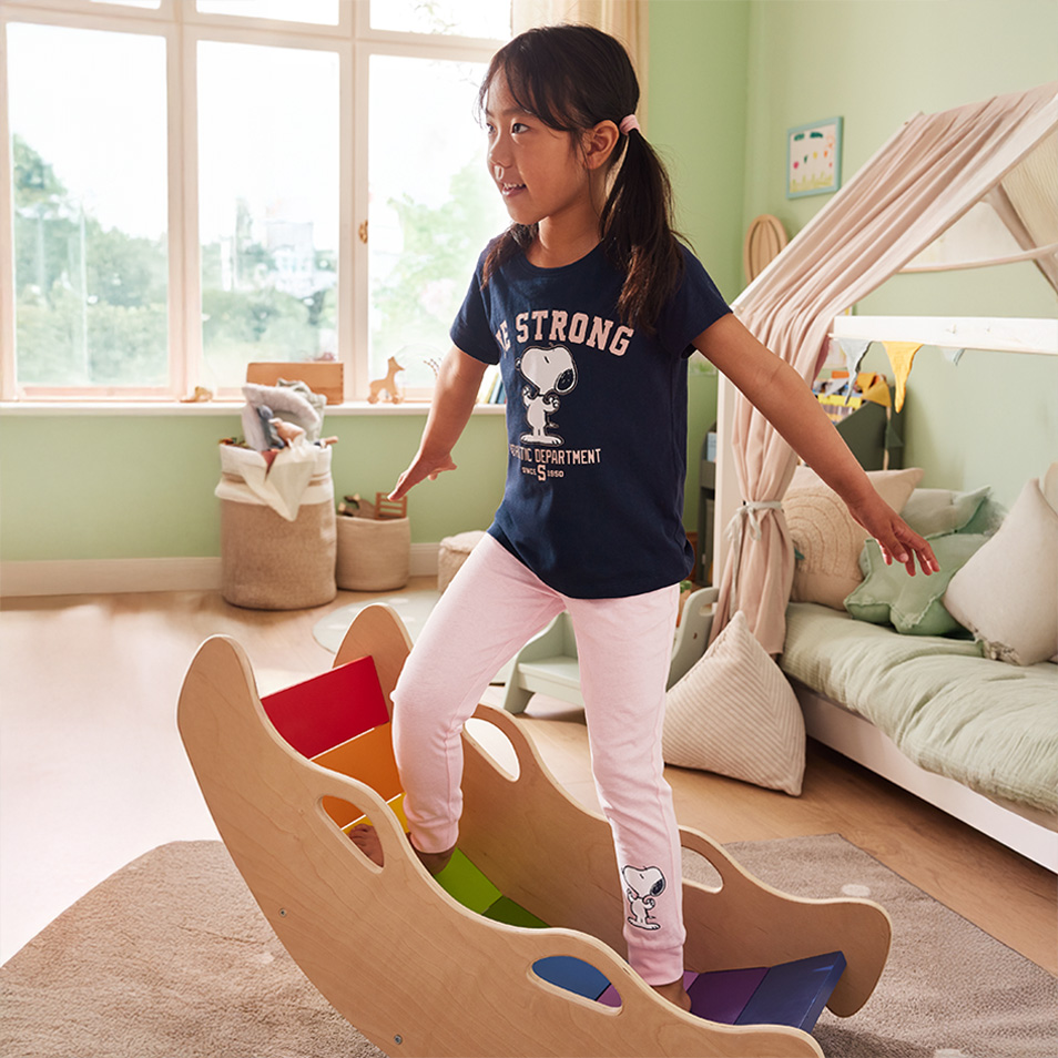 Girl playing on a wooden rainbow-colored rocker, wearing a t-shirt with 'BE STRONG'.