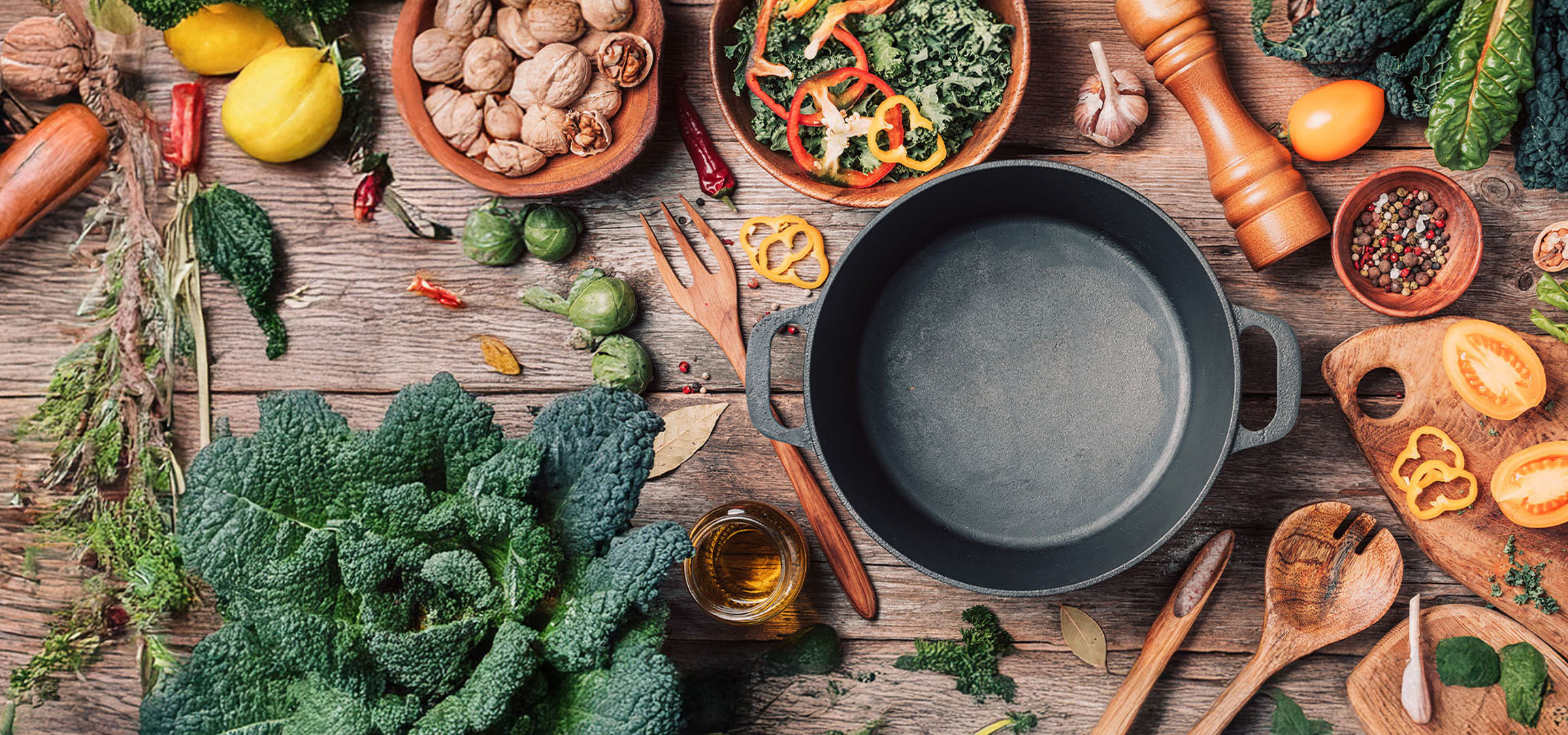 A cast iron pan surrounded by fresh vegetables, nuts, and spices on a wooden table.