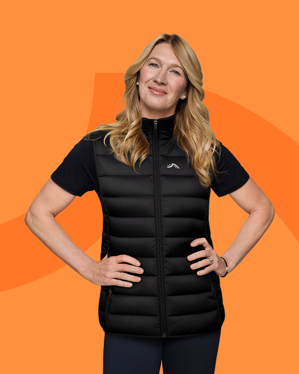 Woman in a black puffer vest and t-shirt, hands on hips, smiling against an orange background.