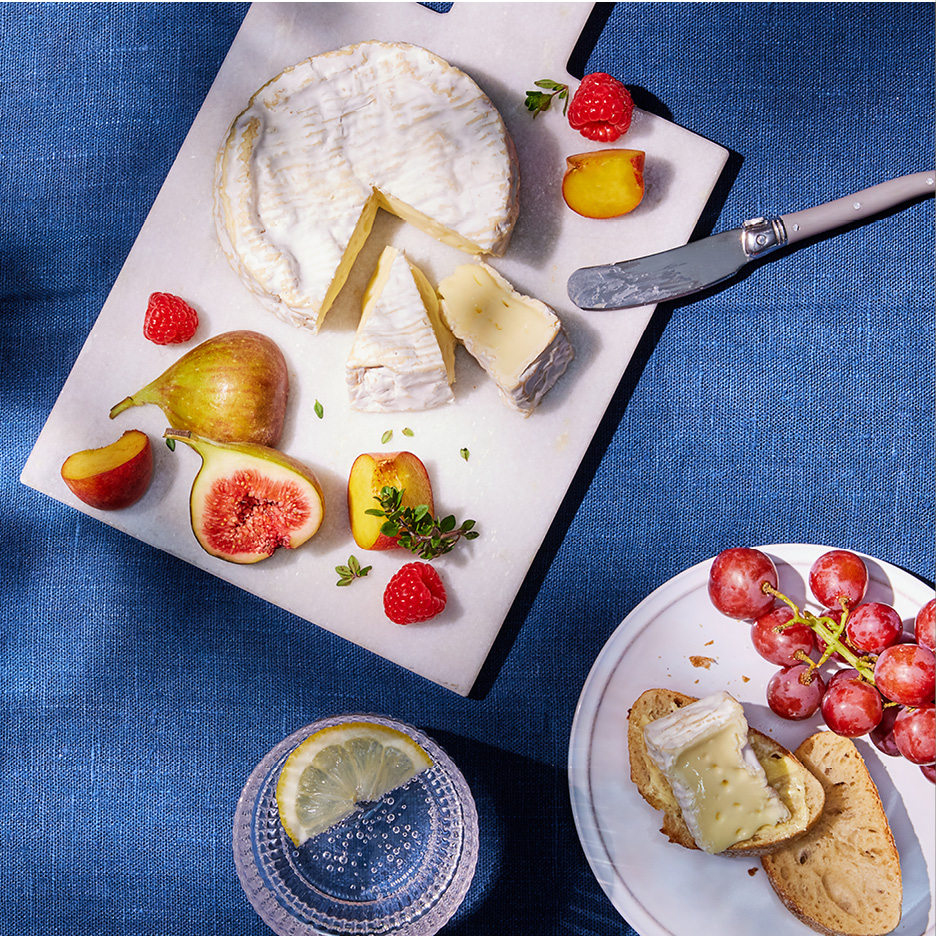 Cheese board with brie, figs, raspberries, and peaches on a blue tablecloth.