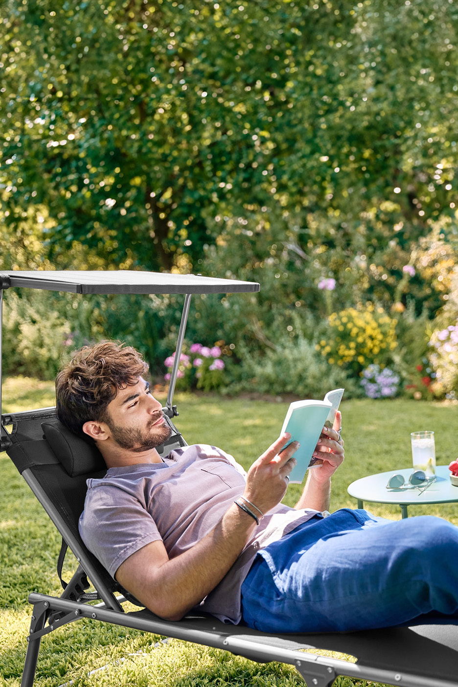 Man relaxing on a garden sun lounger with a sunshade, reading a book in a sunny garden.