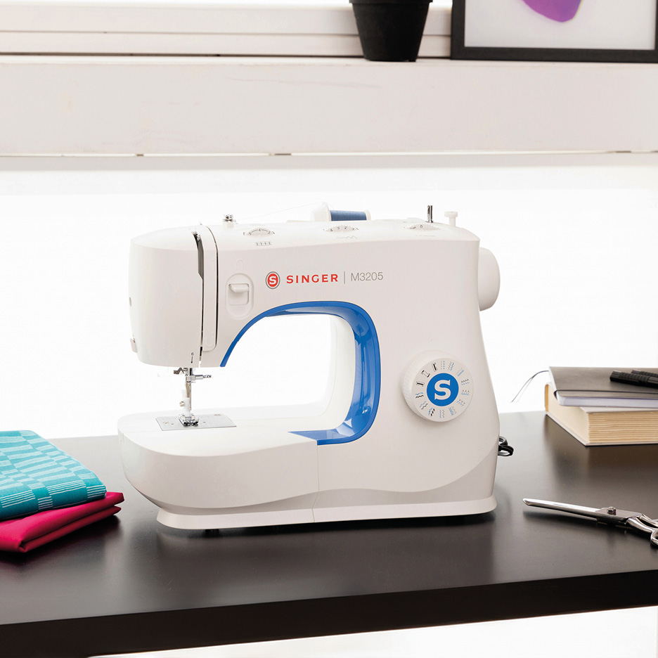 White and blue sewing machine on a dark desk with fabric and notebooks.
