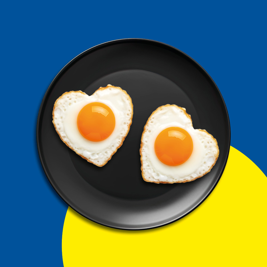 Two heart-shaped fried eggs on a black plate, set against a blue and yellow background.