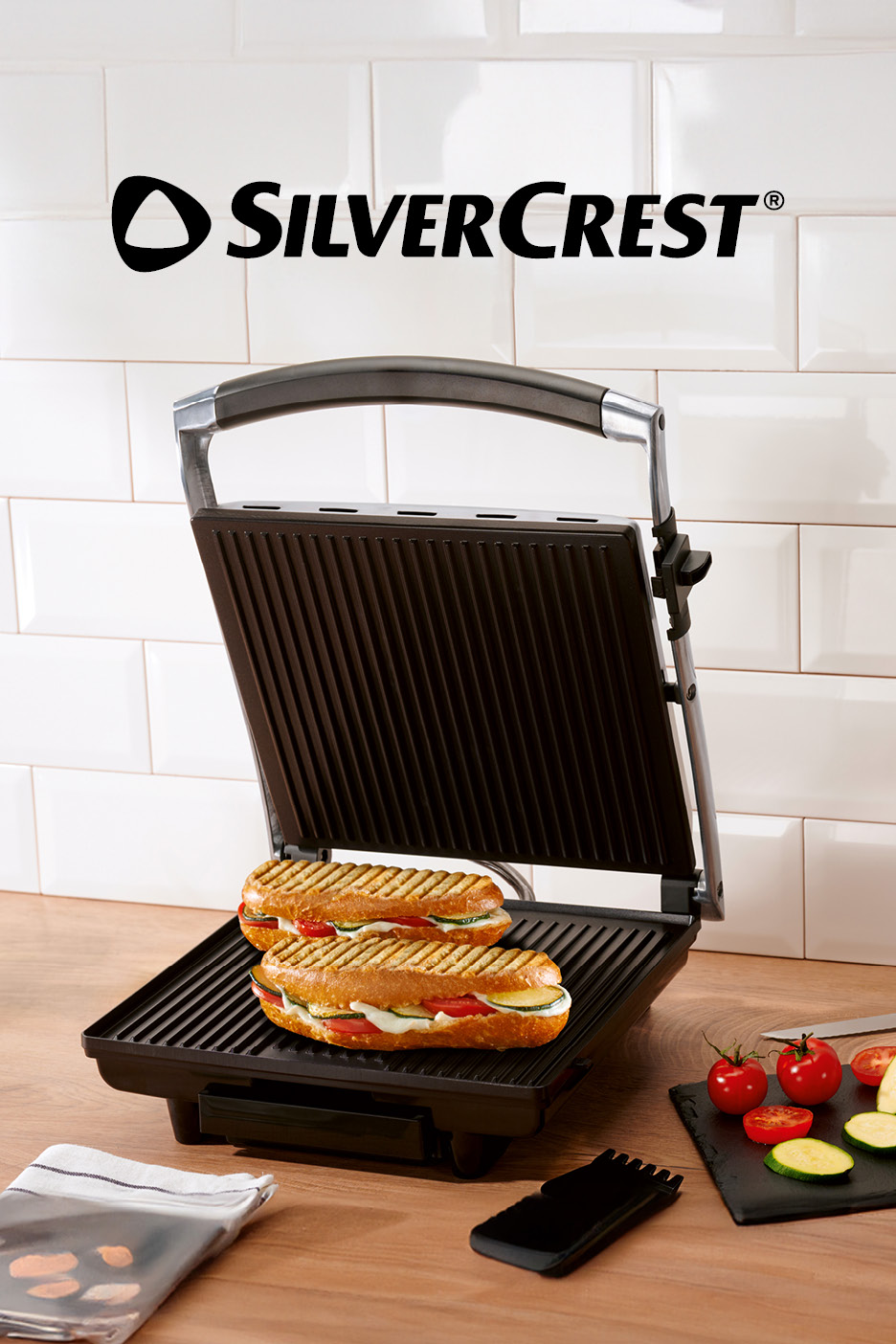 Open grill with two toasted sandwiches, vegetables, and a cleaning tool on a wooden surface.