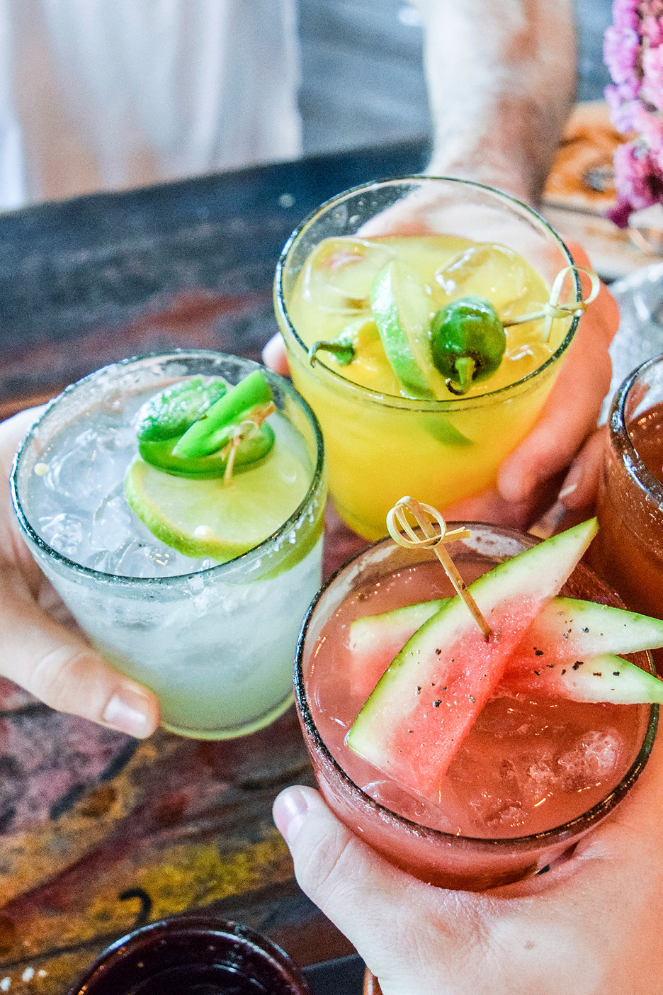 Three hands holding cocktails: a watermelon cocktail, a lime and jalapeño cocktail, and a yellow citrus cocktail.