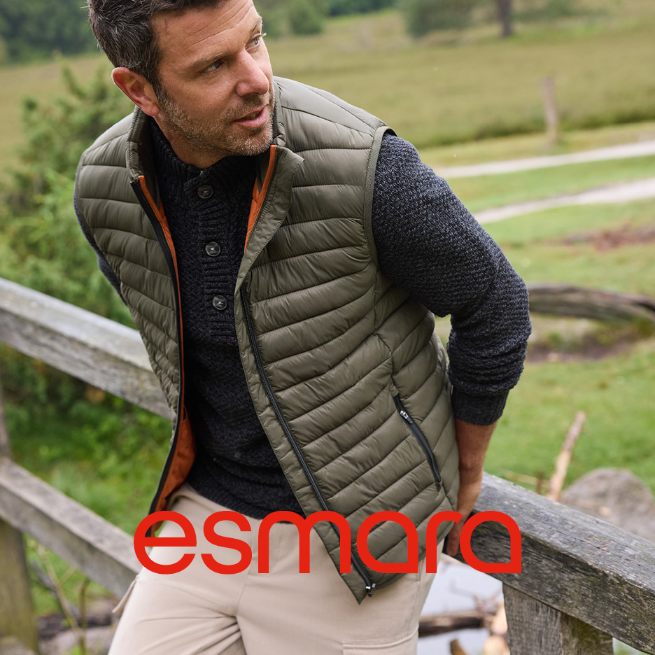 Man in a green quilted vest and dark grey sweater, leaning on a wooden fence outdoors.