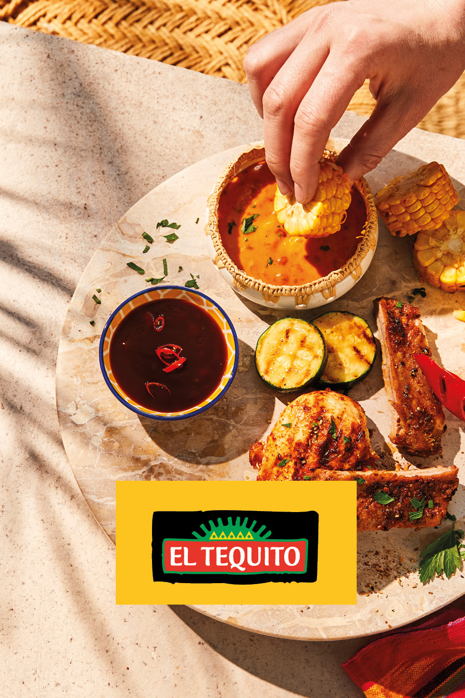 Hand dipping corn into a sauce, with grilled meat, zucchini, and another sauce on a platter.